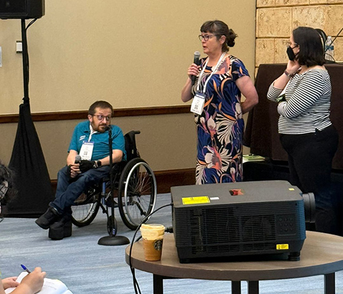 AccessComputing Director Brianna Blaser hosts two speakers with microphones towards the front of a room.