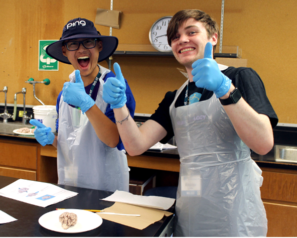 Two students wearing protective lab equipment smile and hold a thumbs up for the camera