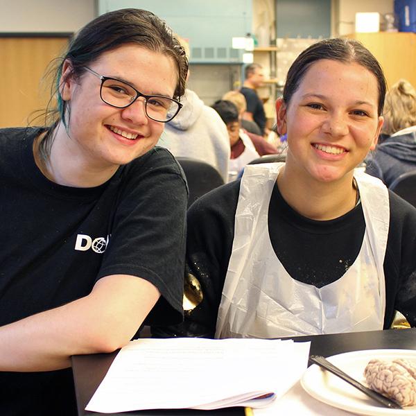 A teaching assistant in a DO-IT shirt and a student in a lab smock are smiling towards the camera in front of a brain dissection lab.