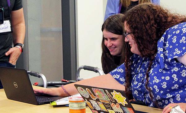 Two individuals look at laptops together, with others standing in the background and an accessibility aid nearby.