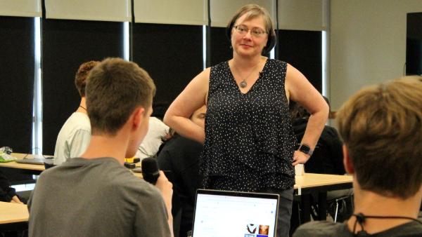  woman standing in a classroom, listening attentively to a student holding a microphone while looking at a laptop screen.