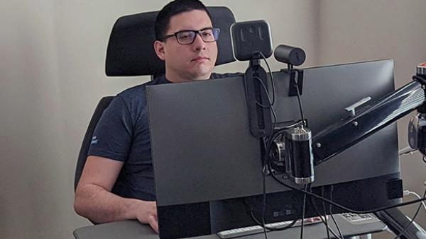 A man sitting in a wheelchair using a large computer setup with multiple monitors and a webcam.