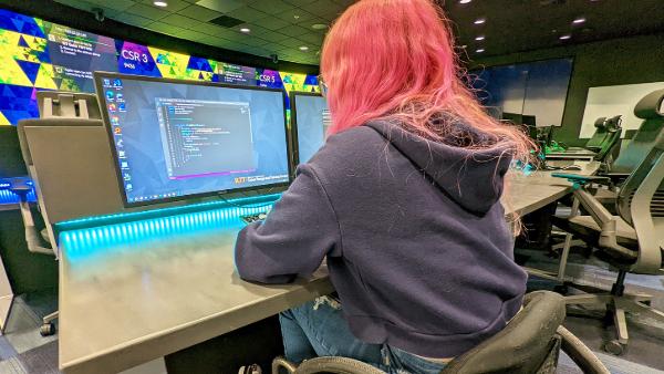 A student sitting at a computer desk in a lab environment, working on coding displayed on the screen.
