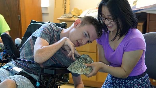A student in a wheelchair closely examining a large rock, while another student holds the rock and assists him.